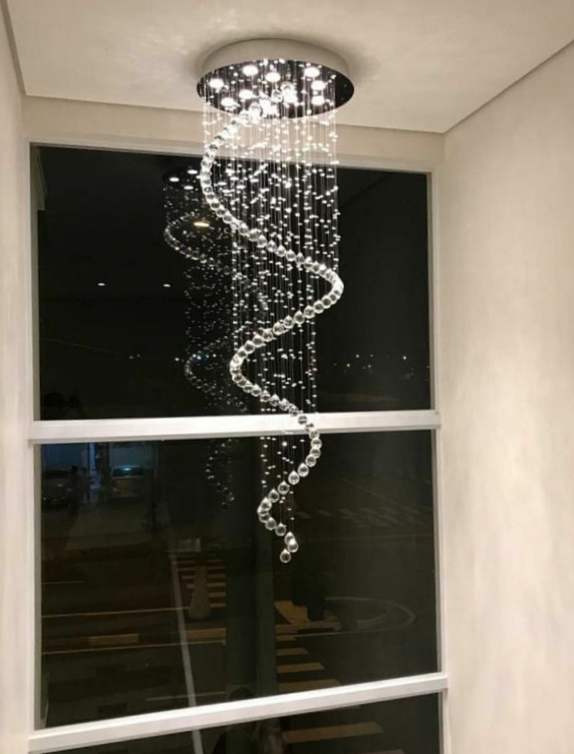 Lustre de cristal modelo "Caracol" da Linha Kria instalado em ambiente com pé direito duplo, com cristais K9 legítimos em espiral decorando sala de estar moderna.